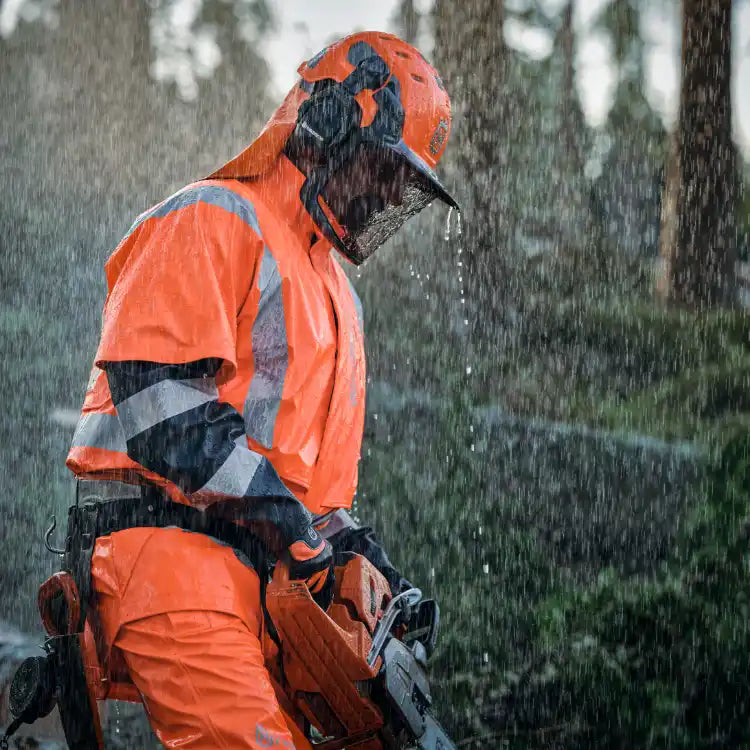 A person in bright orange protective gear holding a chainsaw in the rain.